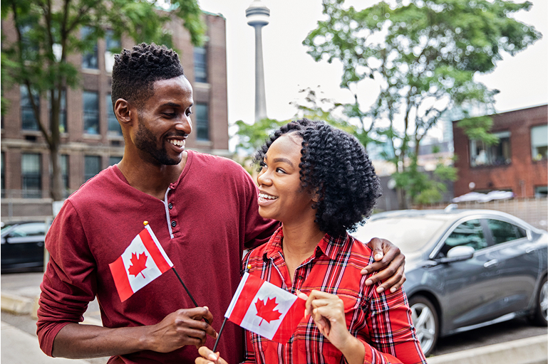 A young couple dressed in red holds Canadian flags in Toronto; the CN Tower can be seen in the background.