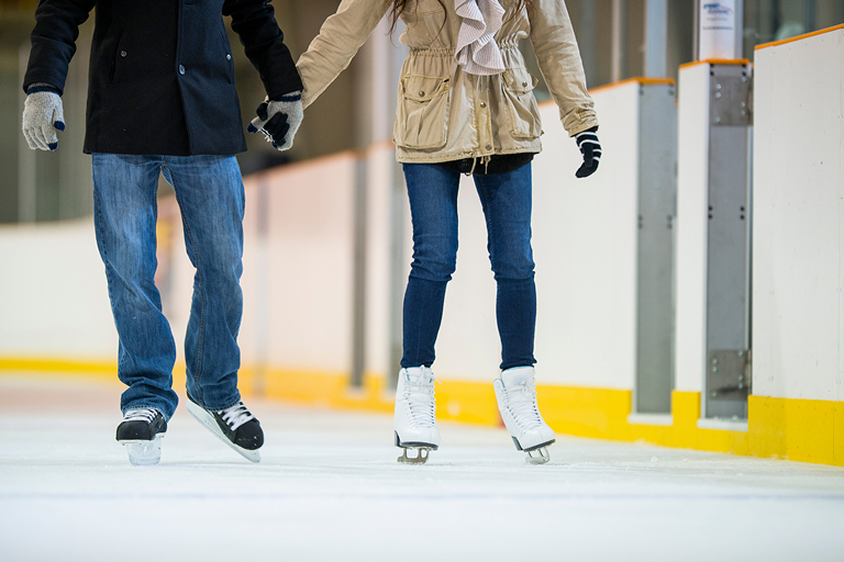 Two people hold hands and skate on an indoor hockey rink.