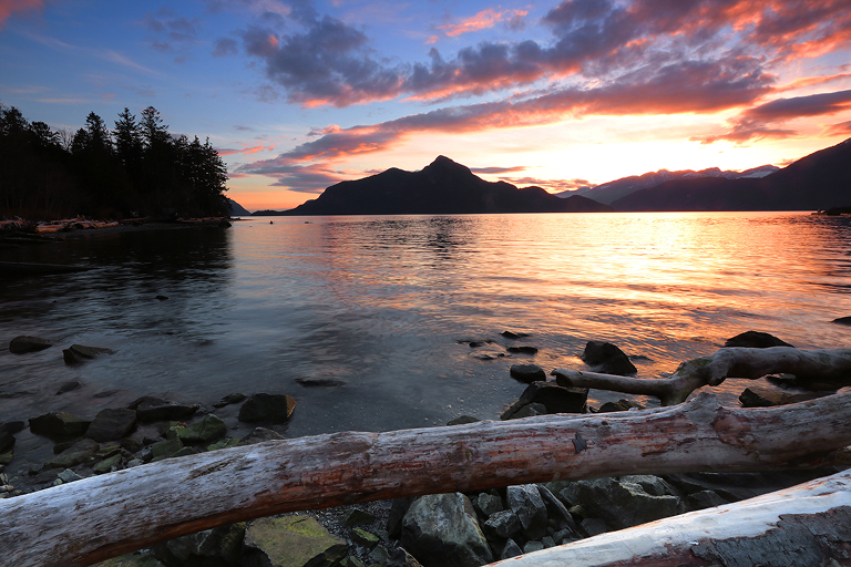 A beach-front photo from British Columbia; the sun sets behind a mountain and reflects off the water.