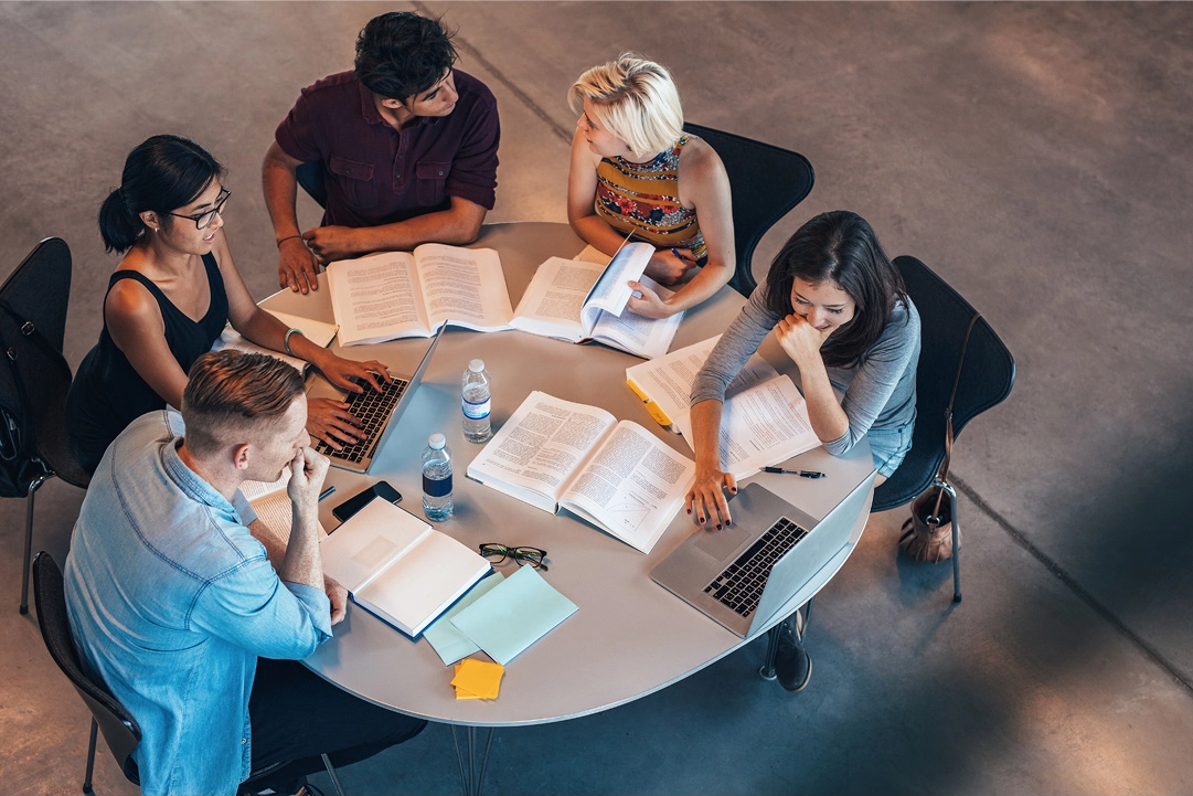 Five young adult students sit at a table and study together.