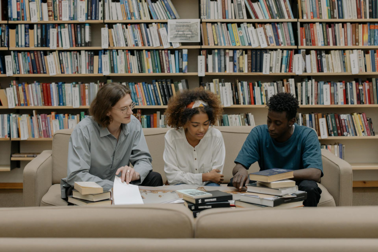 Three people sitting on a couch in a library, reviewing books and documents together, with shelves of books in the background