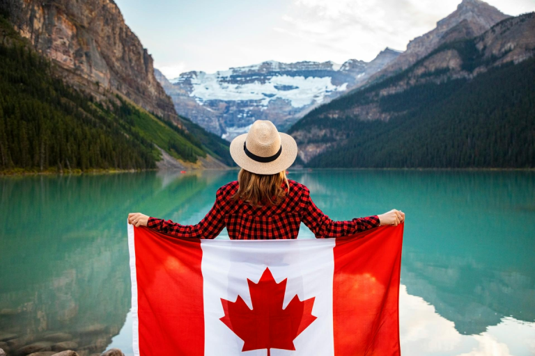 A person standing by a turquoise mountain lake, holding a Canadian flag, with forested slopes and snow-capped peaks in the background