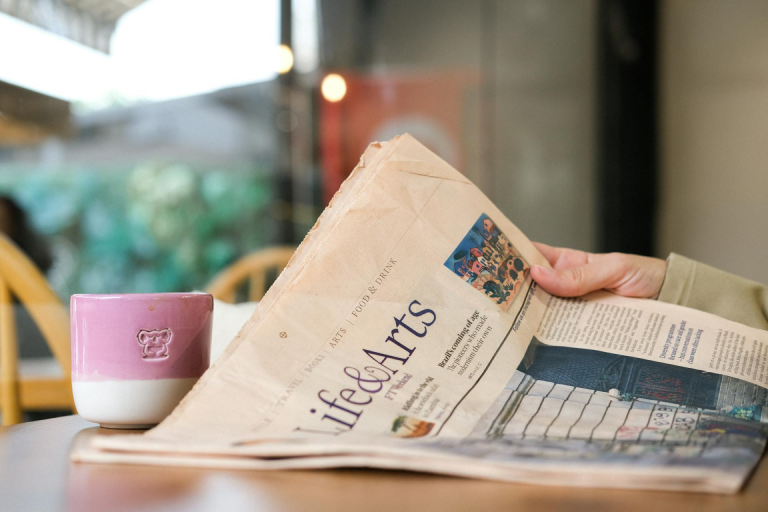 A person holding a newspaper at a café table, with a pink coffee mug sitting beside it