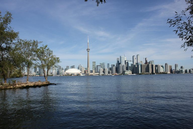 A view of the Toronto skyline across the water, with the CN Tower rising above the city and trees framing the shoreline